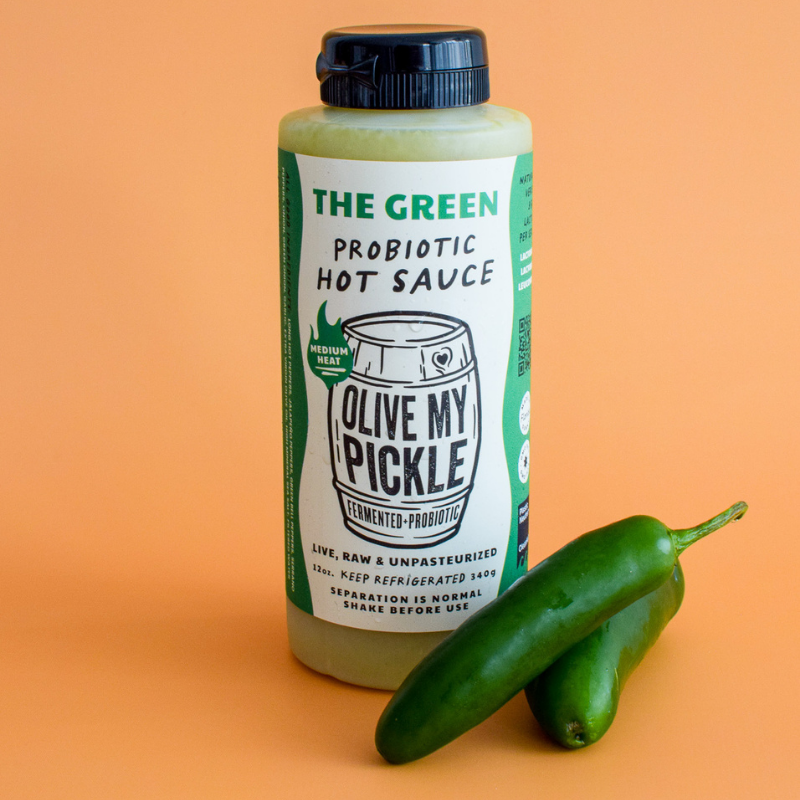 The Green Probiotic Hot Sauce from The hot sauce is next to a pickle on a yellow background