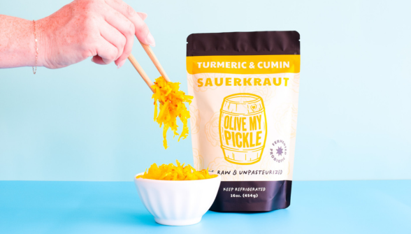 A person's hand holding a jar of pickles over a bowl of pickles. The jar has a label that reads "YUMMY & CUMIN SAUERKRAUT" and "PICKLE" in white text on a yellow background