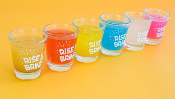 Six Rise and Brine cocktail glasses in different colored drinks arranged on yellow background