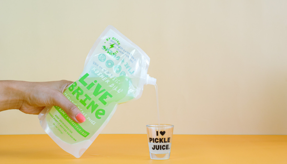 Person pouring green liquid from a bottle into a shot glass. The bottle has a label that reads 'LIVE JUICE' and a person's hand is visible in the background