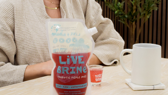 Person in cream sweater holding Live Brine product with white mug and desk on green background